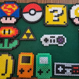 May include: A collection of colorful pixel art designs, including a Superman mushroom, a Pokeball, a question mark block, a mushroom, a flower, a triangle, a controller, a remote, a coin, and two handheld game consoles. The items are arranged on a green surface.