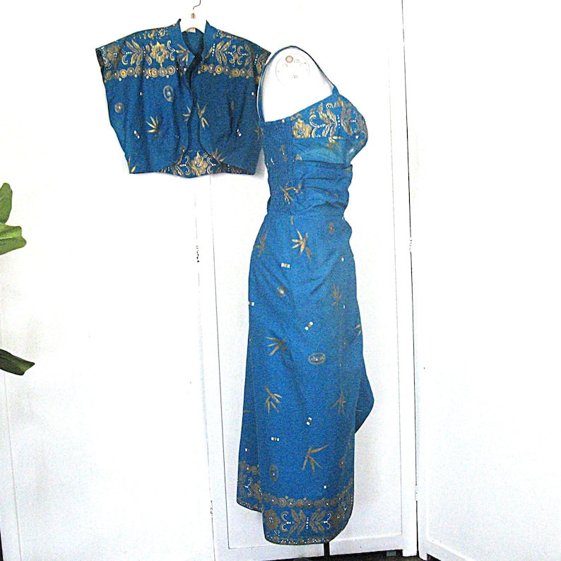 May include: A teal two-piece dress set with gold floral and leaf embroidery. The set comprises a sleeveless dress and a short-sleeved bolero jacket. The dress has a fitted bodice and a flowing skirt. The bolero jacket has a round neckline.