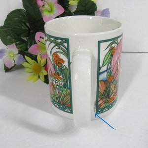 May include: White ceramic mug featuring a pink flamingo, green leaves, and orange flowers. The mug has a green and blue border around the image. The text "mingo" is visible on the mug. The bottom of the mug has the text "8" and "W.S.112" in black.