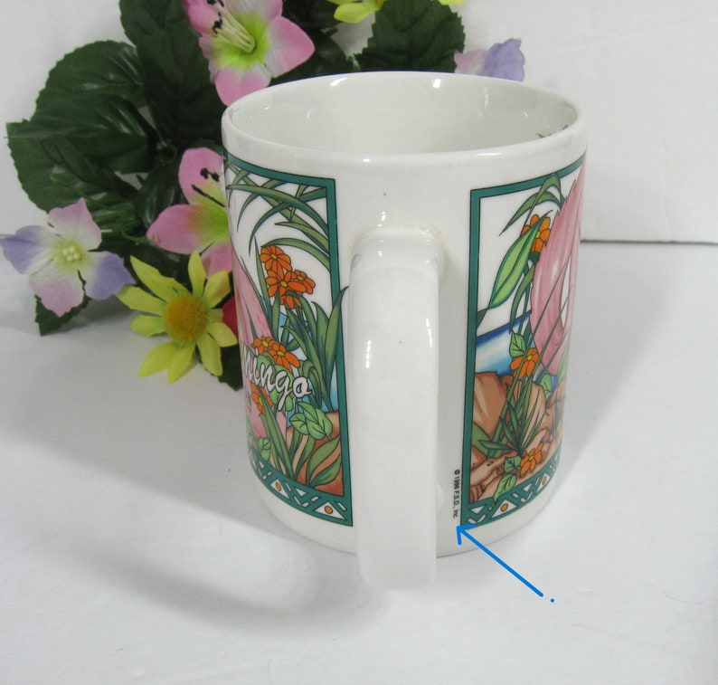 May include: White ceramic mug featuring a pink flamingo, green leaves, and orange flowers. The mug has a green and blue border around the image. The text "mingo" is visible on the mug. The bottom of the mug has the text "8" and "W.S.112" in black.