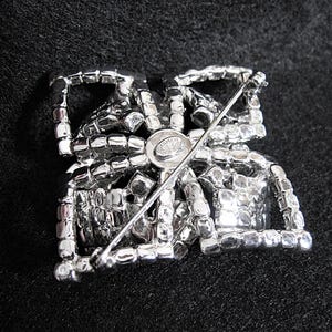May include: The reverse of a silver-toned brooch with a geometric design. The brooch is covered in clear rhinestones and has a pin and catch fastening. The back of the brooch has a small round metal tag with the word "Schumer" printed on it.