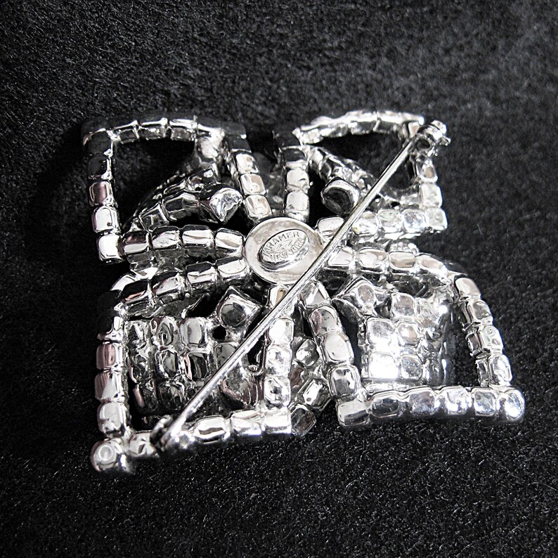May include: The reverse of a silver-toned brooch with a geometric design. The brooch is covered in clear rhinestones and has a pin and catch fastening. The back of the brooch has a small round metal tag with the word "Schumer" printed on it.