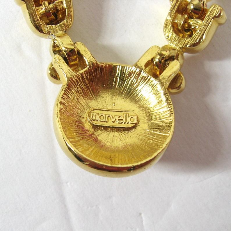 May include: Gold-tone necklace with a round pendant that says "marvella" on the back. The necklace is made of linked metal pieces with small clear stones.