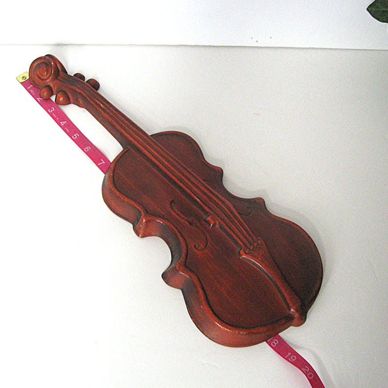 K&ouml;nnte beinhalten: Ein dunkelbraunes, geigenf&ouml;rmiges Wanddekor mit Schnecke, Wirbeln und F-L&ouml;chern. Ein rosa Ma&szlig;band liegt diagonal &uuml;ber dem Objekt und zeigt eine L&auml;nge von &uuml;ber 48 cm von der Schnecke bis zur Unterseite des Instruments an.