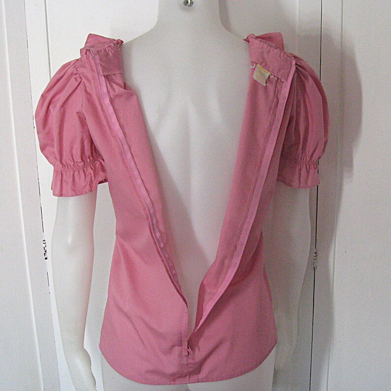 Puede incluir: Una blusa rosa s&oacute;lida con mangas cortas abullonadas con pu&ntilde;os fruncidos y una larga cremallera vertical que se extiende desde el cuello hasta la parte central de la espalda. La prenda se muestra en un maniqu&iacute; con la cremallera trasera abierta.
