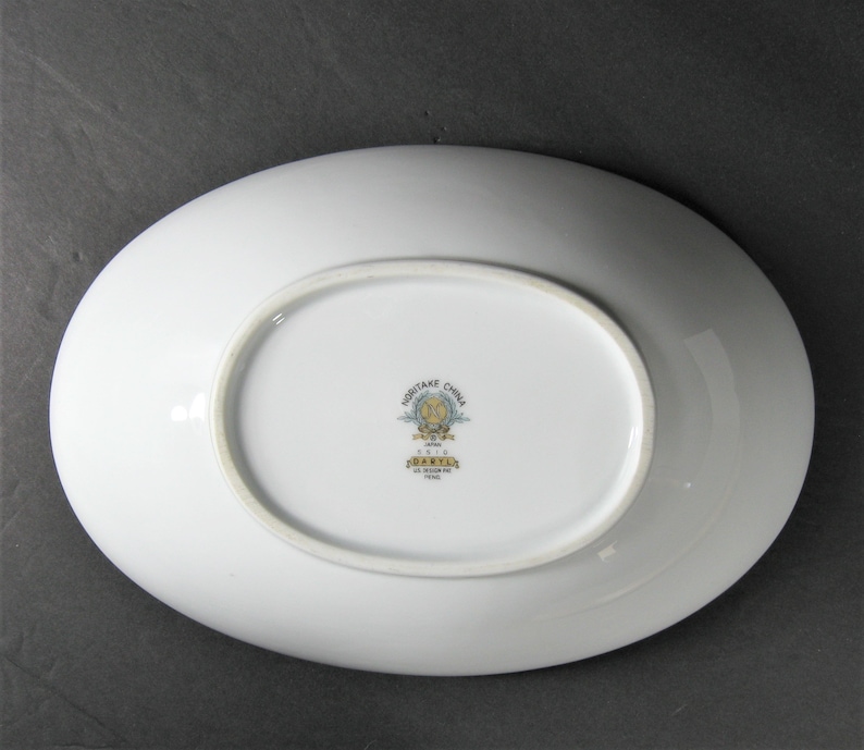 Puede incluir: Plato de servir ovalado blanco con un logotipo de Noritake China en oro y azul. El logotipo incluye el texto "Noritake China" y "Japan" y un escudo con la letra "N".