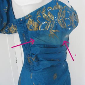 May include: A teal dress with gold floral and bamboo leaf patterns. The dress has a fitted bodice with shirring and a gathered waist. The fabric has a subtle ombre effect. The dress is sleeveless with a scoop neckline.