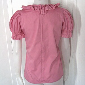 Puede incluir: Una blusa rosa de manga corta con cuello de volantes y mangas abullonadas con pu&ntilde;os el&aacute;sticos. La prenda tiene una costura vertical en el centro de la espalda y se muestra sobre un maniqu&iacute; para exhibir su ajuste y estructura general.