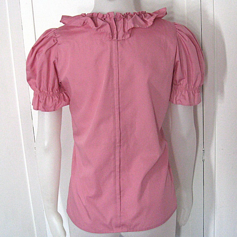 Puede incluir: Una blusa rosa de manga corta con cuello de volantes y mangas abullonadas con pu&ntilde;os el&aacute;sticos. La prenda tiene una costura vertical en el centro de la espalda y se muestra sobre un maniqu&iacute; para exhibir su ajuste y estructura general.