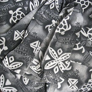 May include: A fabric garment featuring a repeating pattern of white sea turtles and geometric floral shapes set against a dark gray and black background with a subtle marbled texture and visible stitched seams throughout the folded material.