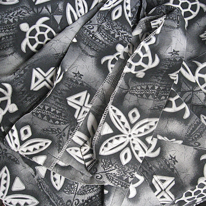 May include: A fabric garment featuring a repeating pattern of white sea turtles and geometric floral shapes set against a dark gray and black background with a subtle marbled texture and visible stitched seams throughout the folded material.