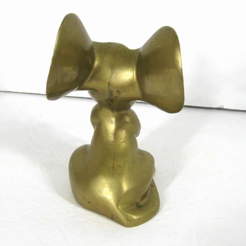 May include: A brass sculpture of a stylized elephant with large, rounded ears. The elephant is sitting on its haunches with its trunk tucked under its body.