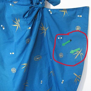 May include: Close-up of blue fabric with gold and white patterns. The fabric has a tie closure and features bamboo-like designs, geometric shapes, and arrow graphics. The fabric is likely part of a garment, possibly a kimono.