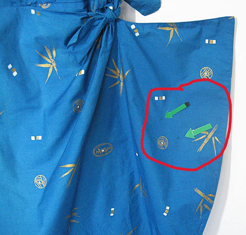 May include: Close-up of blue fabric with gold and white patterns. The fabric has a tie closure and features bamboo-like designs, geometric shapes, and arrow graphics. The fabric is likely part of a garment, possibly a kimono.
