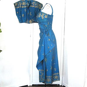 May include: A teal two-piece dress set with gold floral and geometric patterns. The set includes a sleeveless dress with a wrap skirt and a matching short-sleeved bolero jacket. The dress has spaghetti straps and a fitted bodice. The fabric appears to be a lightweight material.