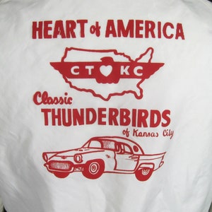 Pode incluir: Uma camiseta branca com uma estampa gr&aacute;fica vermelha e branca. A estampa apresenta um mapa estilizado dos Estados Unidos com o texto "HEART of AMERICA" acima do mapa e "Classic THUNDERBIRDS of Kansas City" abaixo do mapa. O mapa tem um cora&ccedil;&atilde;o vermelho no centro com o texto "CT KC" acima do cora&ccedil;&atilde;o.