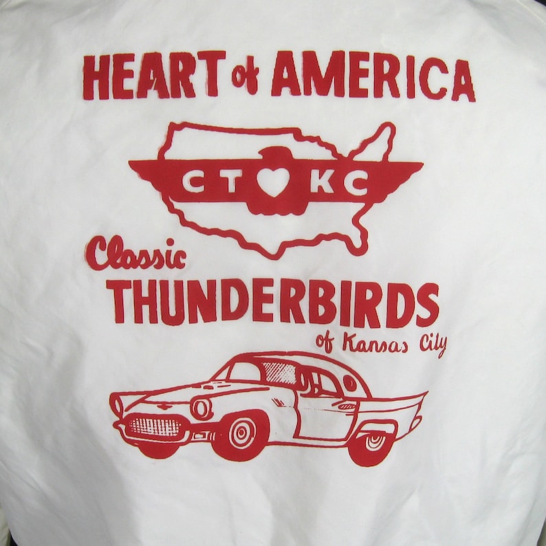 Pode incluir: Uma camiseta branca com uma estampa gr&aacute;fica vermelha e branca. A estampa apresenta um mapa estilizado dos Estados Unidos com o texto "HEART of AMERICA" acima do mapa e "Classic THUNDERBIRDS of Kansas City" abaixo do mapa. O mapa tem um cora&ccedil;&atilde;o vermelho no centro com o texto "CT KC" acima do cora&ccedil;&atilde;o.