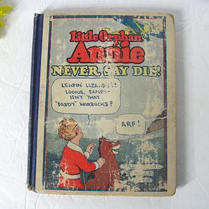 Pu&ograve; includere: Una copertina di libro vintage con un'illustrazione a fumetti di Little Orphan Annie e il suo cane Sandy. Il titolo del libro &egrave; "Little Orphan Annie NEVER SAY DIE!" con il testo "LEAPIN' LIZARDS!! LOOKIE, SANDY - ISN'T THAT "DADDY" WARBUCKS?" in una nuvola di dialogo sopra Annie. Sandy dice "ARF!" in una nuvola di dialogo. Il libro &egrave; pubblicato da Cupples & Leon Co., New York.