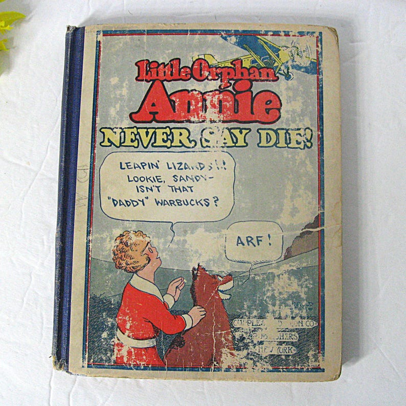 Pu&ograve; includere: Una copertina di libro vintage con un'illustrazione a fumetti di Little Orphan Annie e il suo cane Sandy. Il titolo del libro &egrave; "Little Orphan Annie NEVER SAY DIE!" con il testo "LEAPIN' LIZARDS!! LOOKIE, SANDY - ISN'T THAT "DADDY" WARBUCKS?" in una nuvola di dialogo sopra Annie. Sandy dice "ARF!" in una nuvola di dialogo. Il libro &egrave; pubblicato da Cupples & Leon Co., New York.