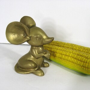 May include: A brass figurine of a mouse with large ears. The mouse is sitting on its hind legs with its front paws together. The mouse is facing to the right. The figurine is in focus, while a yellow corn cob is out of focus in the background.