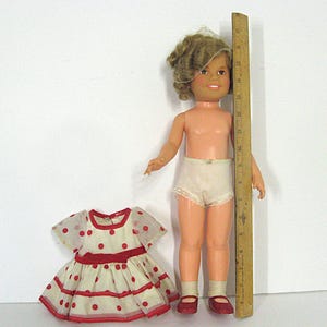 May include: A doll with blonde hair, wearing white lace-trimmed underwear and red shoes, stands next to a wooden ruler measuring approximately 18 inches. Beside the doll is a white dress with red polka dots and a red waistband, short sleeves, and trim.