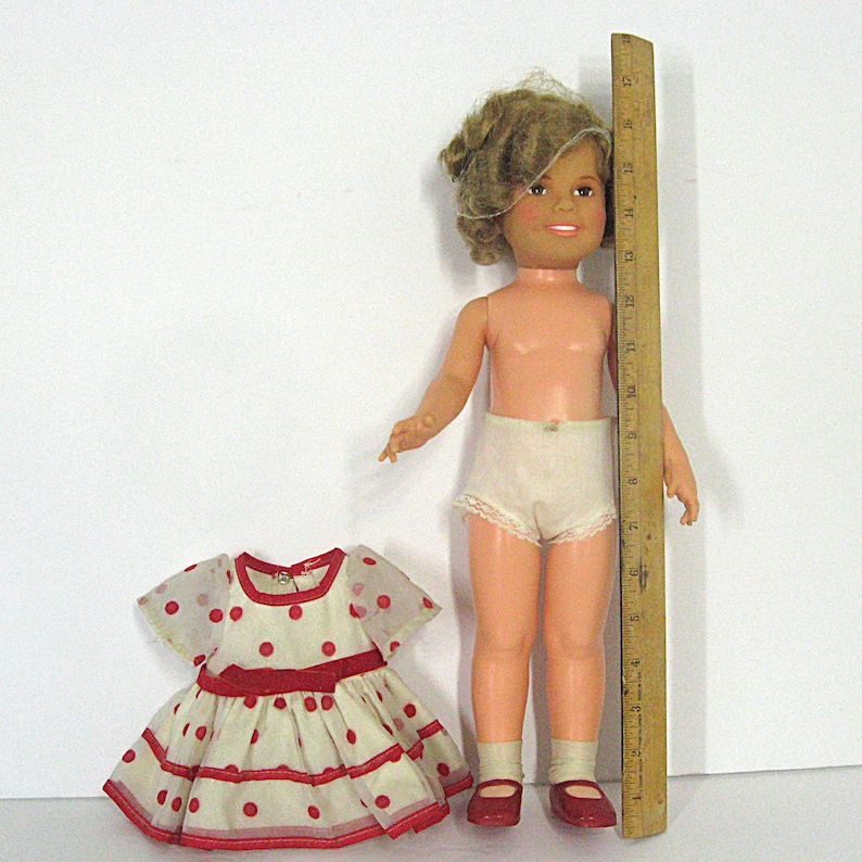 May include: A doll with blonde hair, wearing white lace-trimmed underwear and red shoes, stands next to a wooden ruler measuring approximately 18 inches. Beside the doll is a white dress with red polka dots and a red waistband, short sleeves, and trim.
