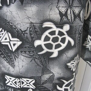 May include: A short-sleeved button-down shirt in shades of black, gray, and white. The fabric features a batik-style pattern with white sea turtles and geometric tribal motifs. A single chest pocket is integrated into the overall print design.