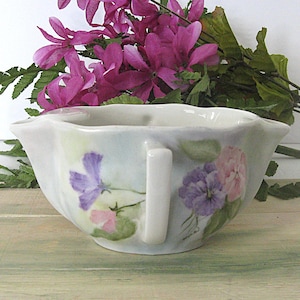 May include: A white porcelain gravy boat with a handle, adorned with hand-painted purple and pink floral designs. The boat has a scalloped rim and is set against a backdrop of pink flowers and green foliage.