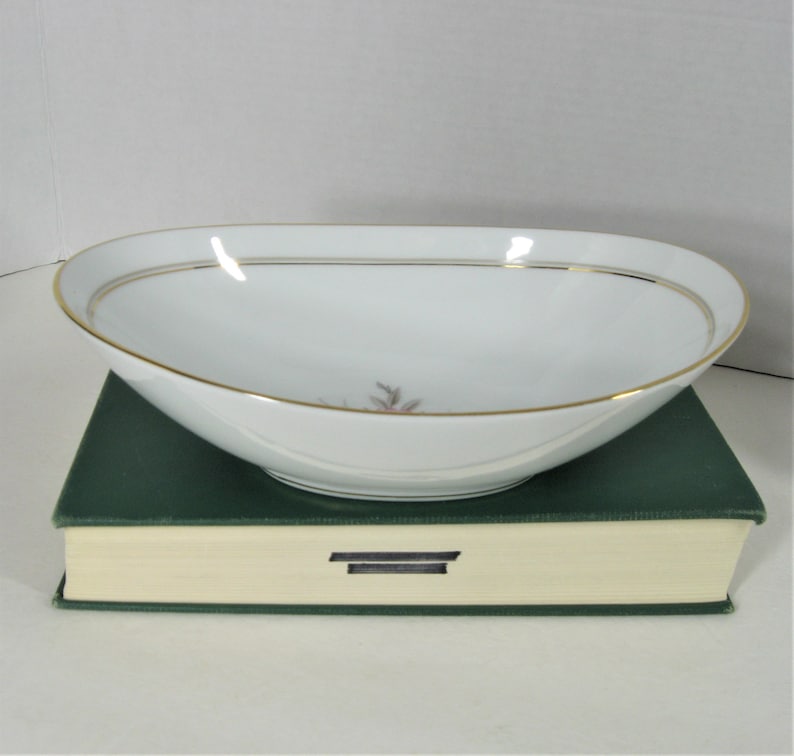 Puede incluir: Taz&oacute;n ovalado blanco con borde dorado y dise&ntilde;o floral. El taz&oacute;n est&aacute; sobre un libro verde.