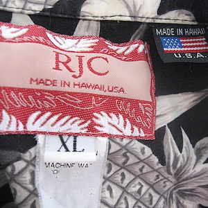 K&ouml;nnte beinhalten: Ein schwarzes Hemd mit wei&szlig;em Ananas-Druck und einem roten RJC-Markenetikett mit der Aufschrift Made in Hawaii, USA. Ein kleineres schwarzes Etikett zeigt den Text Made in Hawaii U.S.A. mit einer US-Flagge. Ein wei&szlig;es Etikett zeigt die Gr&ouml;&szlig;e XL.