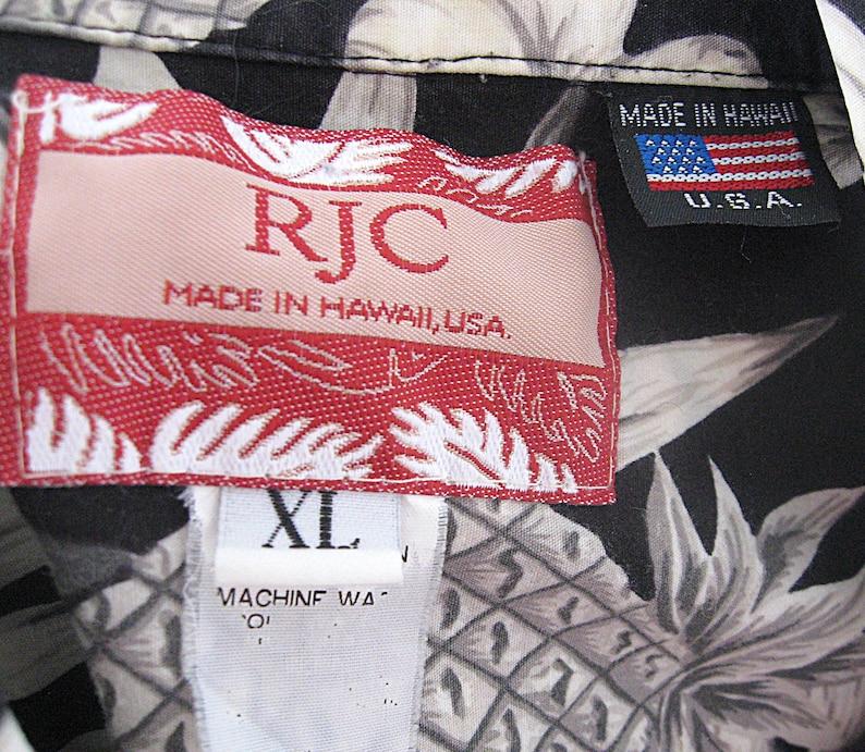 K&ouml;nnte beinhalten: Ein schwarzes Hemd mit wei&szlig;em Ananas-Druck und einem roten RJC-Markenetikett mit der Aufschrift Made in Hawaii, USA. Ein kleineres schwarzes Etikett zeigt den Text Made in Hawaii U.S.A. mit einer US-Flagge. Ein wei&szlig;es Etikett zeigt die Gr&ouml;&szlig;e XL.