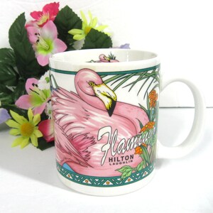 May include: White ceramic mug featuring a pink flamingo design. The mug has a green border and the text "Flamingos Hilton Laughlin" printed on it.