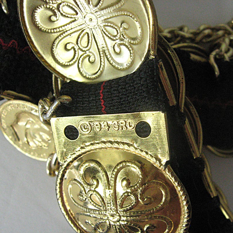 May include: Close-up of a gold-toned belt with ornate circular medallions and a rectangular clasp. The belt is attached to a black fabric base with red stitching. The clasp is stamped with "&copy;94 GRD".
