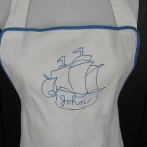 May include: White apron with blue trim and a blue embroidered ship with the name "John" underneath.