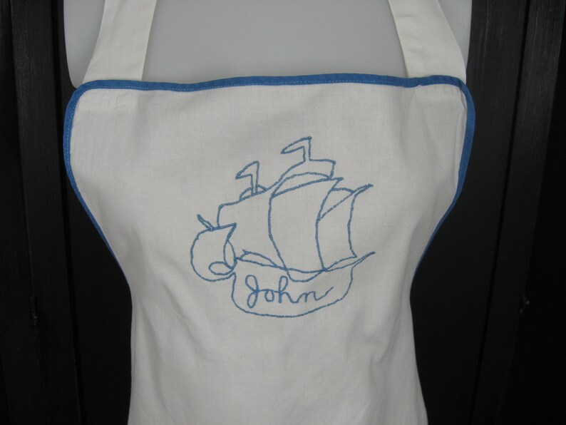May include: White apron with blue trim and a blue embroidered ship with the name "John" underneath.