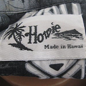 May include: A white rectangular clothing label with black text and graphics stitched onto a black and gray patterned fabric. The label features the word Howie, a palm tree illustration, a mountain graphic, and the text Made in Hawaii.