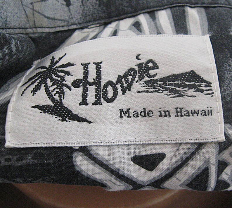 May include: A white rectangular clothing label with black text and graphics stitched onto a black and gray patterned fabric. The label features the word Howie, a palm tree illustration, a mountain graphic, and the text Made in Hawaii.