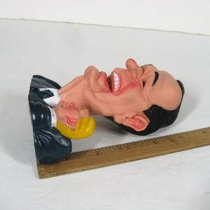 May include: A caricature figurine of two men, one with a large head and a wide open mouth, the other with a smaller head and a closed mouth. The figurine is made of plastic and is painted in realistic colors. The figurine is lying on its side, with the larger head facing the camera.