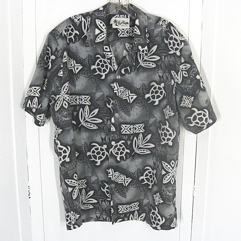 May include: A short-sleeved button-down shirt in shades of gray, black, and white. The fabric features a repeating pattern of sea turtles, tropical leaves, and geometric tribal motifs. The shirt has a collar and a front button closure.