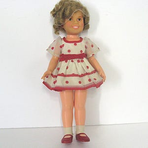 May include: A doll with light brown curly hair and a smiling expression. The doll wears a cream-colored dress featuring red polka dots, short sheer sleeves, and a red ribbon waistband. It also wears light-colored socks and red shoes.