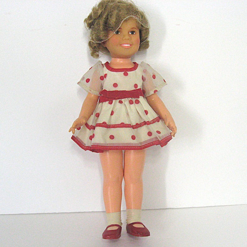 May include: A doll with light brown curly hair and a smiling expression. The doll wears a cream-colored dress featuring red polka dots, short sheer sleeves, and a red ribbon waistband. It also wears light-colored socks and red shoes.