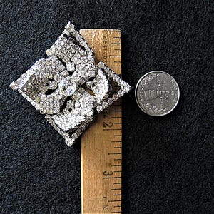 May include: A large, silver-toned, rhinestone brooch in the shape of a square with a floral design. The brooch is approximately 5 cm wide and is next to a UK 20 pence coin for size comparison.