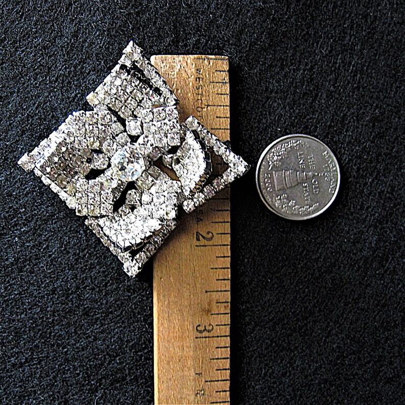 May include: A large, silver-toned, rhinestone brooch in the shape of a square with a floral design. The brooch is approximately 5 cm wide and is next to a UK 20 pence coin for size comparison.