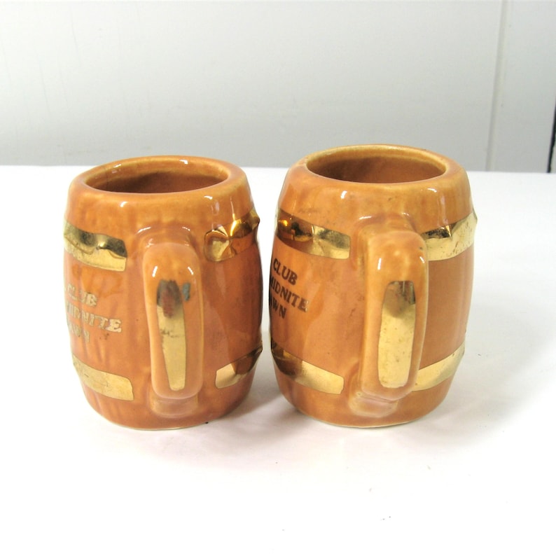 Op de afbeelding: Twee kleine bruine keramische bekers in de vorm van vaten met gouden versieringen en de tekst "The Club Midnite Inn" erop gedrukt.