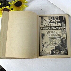 Pu&ograve; includere: Un libro aperto con un'illustrazione in bianco e nero di una ragazza e un cane sulla pagina destra. L'illustrazione &egrave; tratta dalla striscia a fumetti "Little Orphan Annie" e il testo "Never Say Die!" &egrave; sopra l'illustrazione. La pagina sinistra &egrave; vuota.