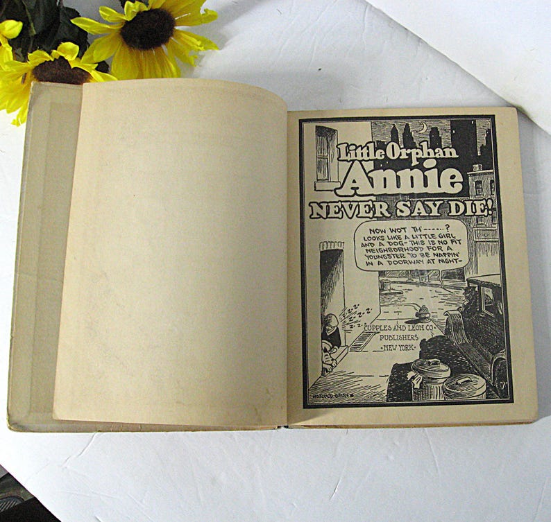 Pu&ograve; includere: Un libro aperto con un'illustrazione in bianco e nero di una ragazza e un cane sulla pagina destra. L'illustrazione &egrave; tratta dalla striscia a fumetti "Little Orphan Annie" e il testo "Never Say Die!" &egrave; sopra l'illustrazione. La pagina sinistra &egrave; vuota.