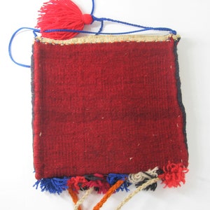 May include: A small, rectangular, woven bag with a deep red central field. The bag has a blue drawstring and a red tassel at the top. The edges are trimmed with a gold-colored material and the bottom has colorful tassels.