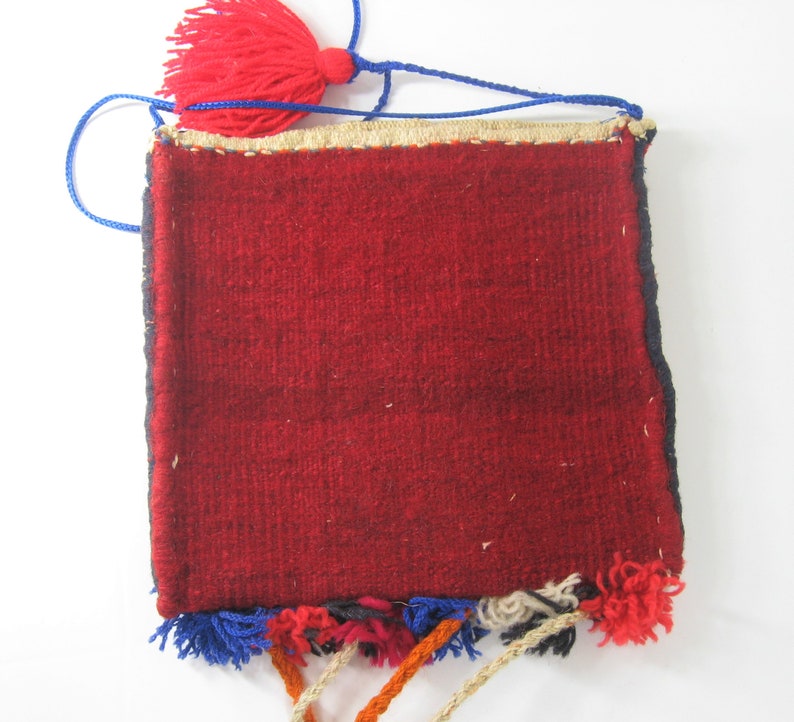 May include: A small, rectangular, woven bag with a deep red central field. The bag has a blue drawstring and a red tassel at the top. The edges are trimmed with a gold-colored material and the bottom has colorful tassels.