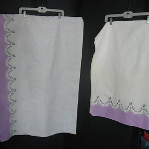May include: Two white linen towels with a lavender border and scalloped edge. The border is adorned with embroidered floral details in shades of green and blue. The towels are hanging on white plastic hangers.