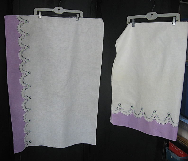 May include: Two white linen towels with a lavender border and scalloped edge. The border is adorned with embroidered floral details in shades of green and blue. The towels are hanging on white plastic hangers.